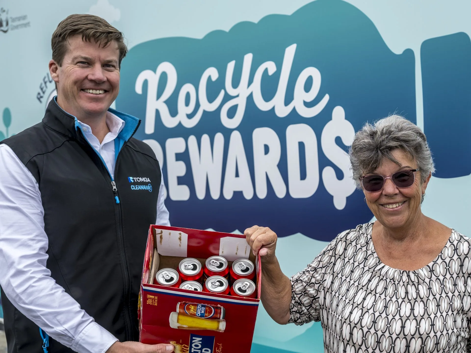 Picture of two people standing in front of a Recycle Rewards reverse vending machine holding a box of empty used aluminium cans.