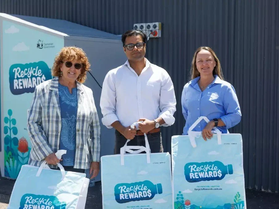 Three people holding reusable bags with Recycle Rewards logos on the front standing in front of a Recycle Rewards reverse vending machine.