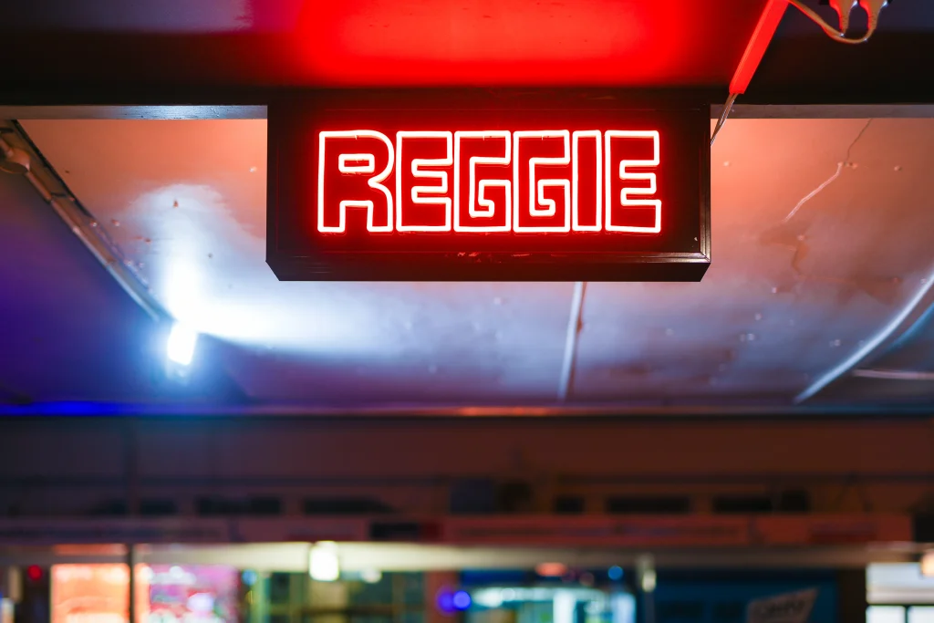 A7403306 1 - TOMRA Cleanaway Red neon sign reading'REGGAE' mounted in a dark ceiling area of a nightclub/bar with blue and purple lighting around it