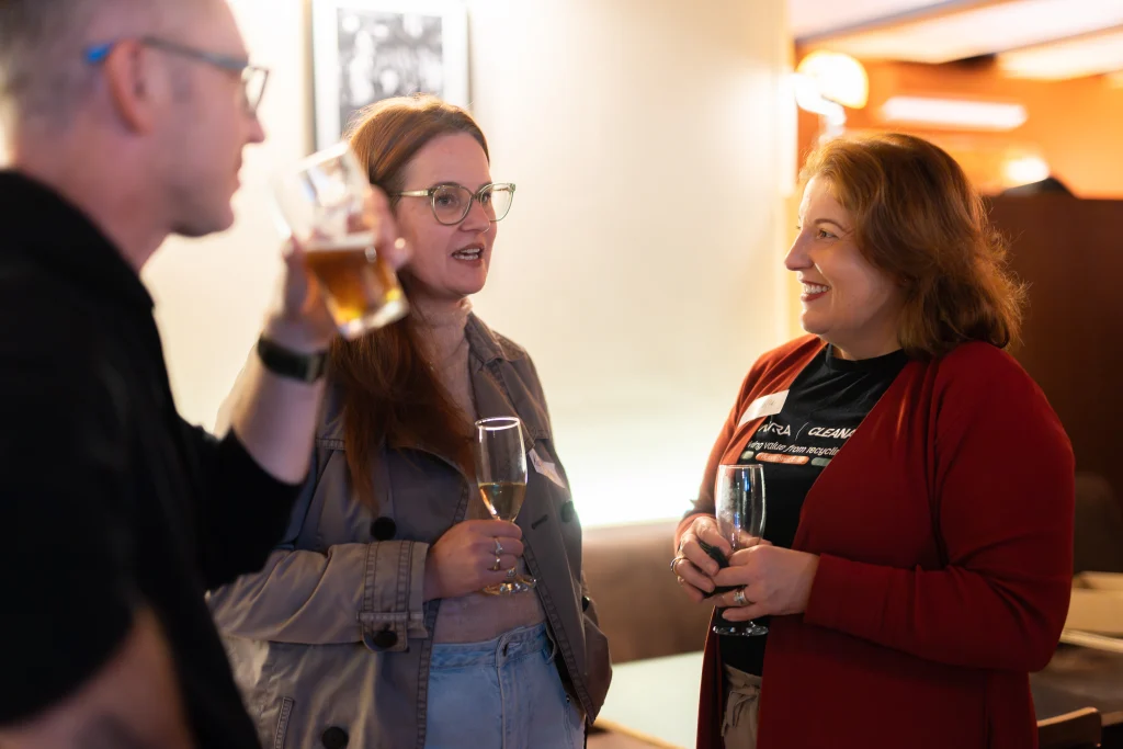 A7403367 1 - TOMRA Cleanaway Three adults conversing at a bar, each holding a drink and smiling at one another.