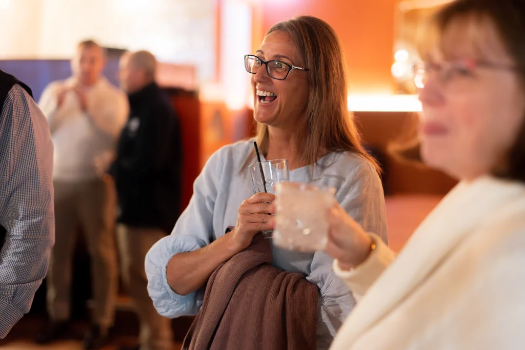 A7403402 1 - TOMRA Cleanaway Woman with glasses laughing while holding a glass with a straw at a social gathering; blurred partygoers in the background.
