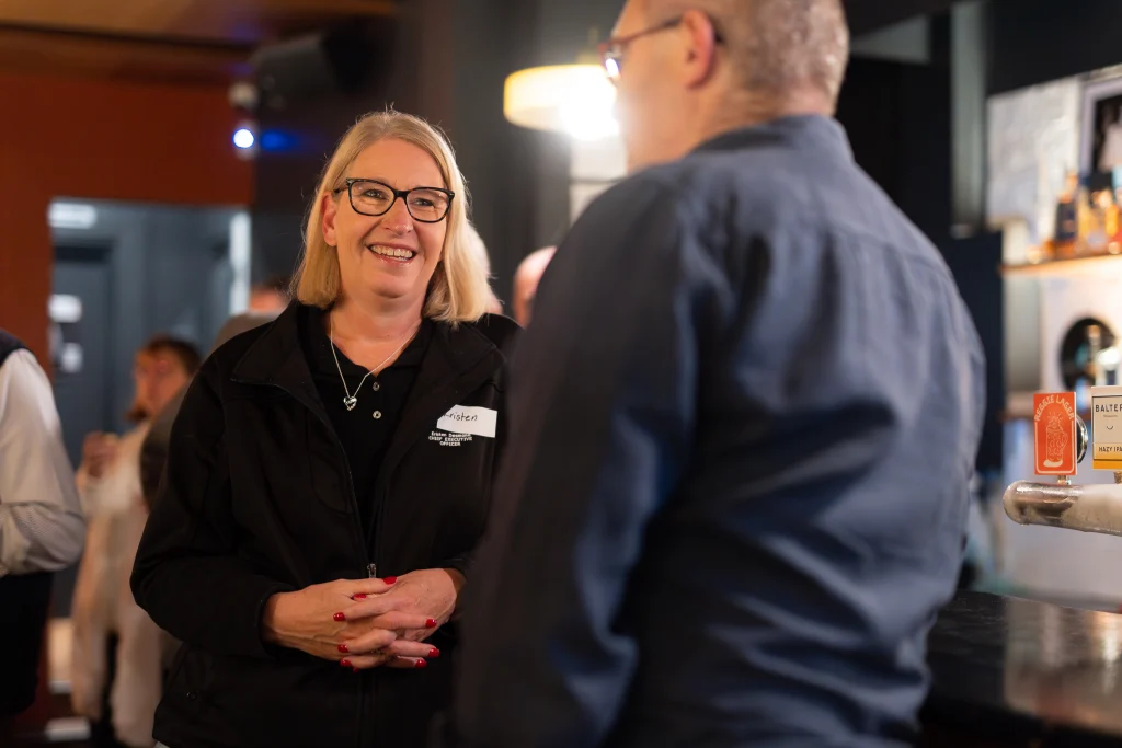A7403414 1 - TOMRA Cleanaway Smiling woman with blonde hair and glasses talking to a man at a bar or counter, wearing a black jacket and name tag.