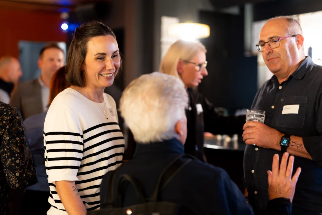 A7403474 1 - TOMRA Cleanaway Smiling woman in a striped shirt chats with a man wearing glasses at a social gathering; others blurred in the background.