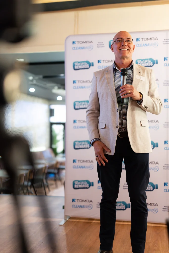 A7403539 1 - TOMRA Cleanaway Smiling man in a beige blazer speaks into a microphone at a branded backdrop backdrop.