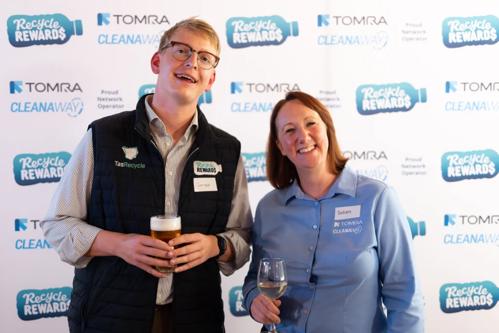A7403568 1 - TOMRA Cleanaway Two smiling adults at a Recycle Rewards event, holding drink glasses, with a TOMRA backdrop behind them.