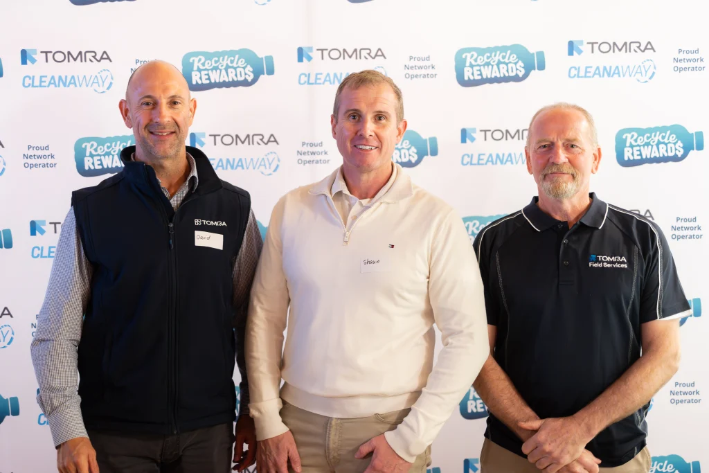 A7403590 1 - TOMRA Cleanaway Three men posing for a photo at a TOMRA recycling event, backdrop shows ‘Recycle Rewards’ logos; left and center name tags read David and Shaun, respectively.