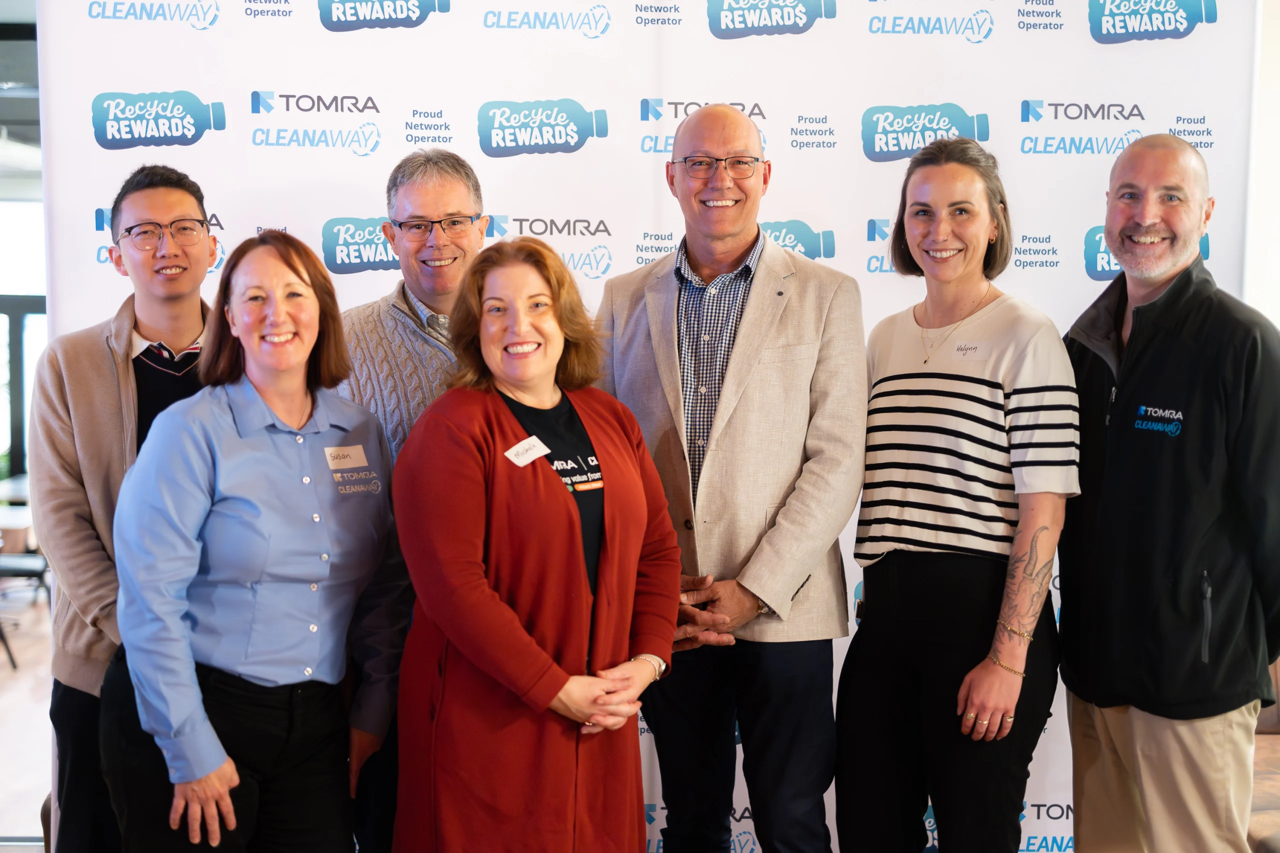 Six people posing for a group photo in front of a branded backdrop featuring Recycle Rewards and Tomra logos; several wear name tags.