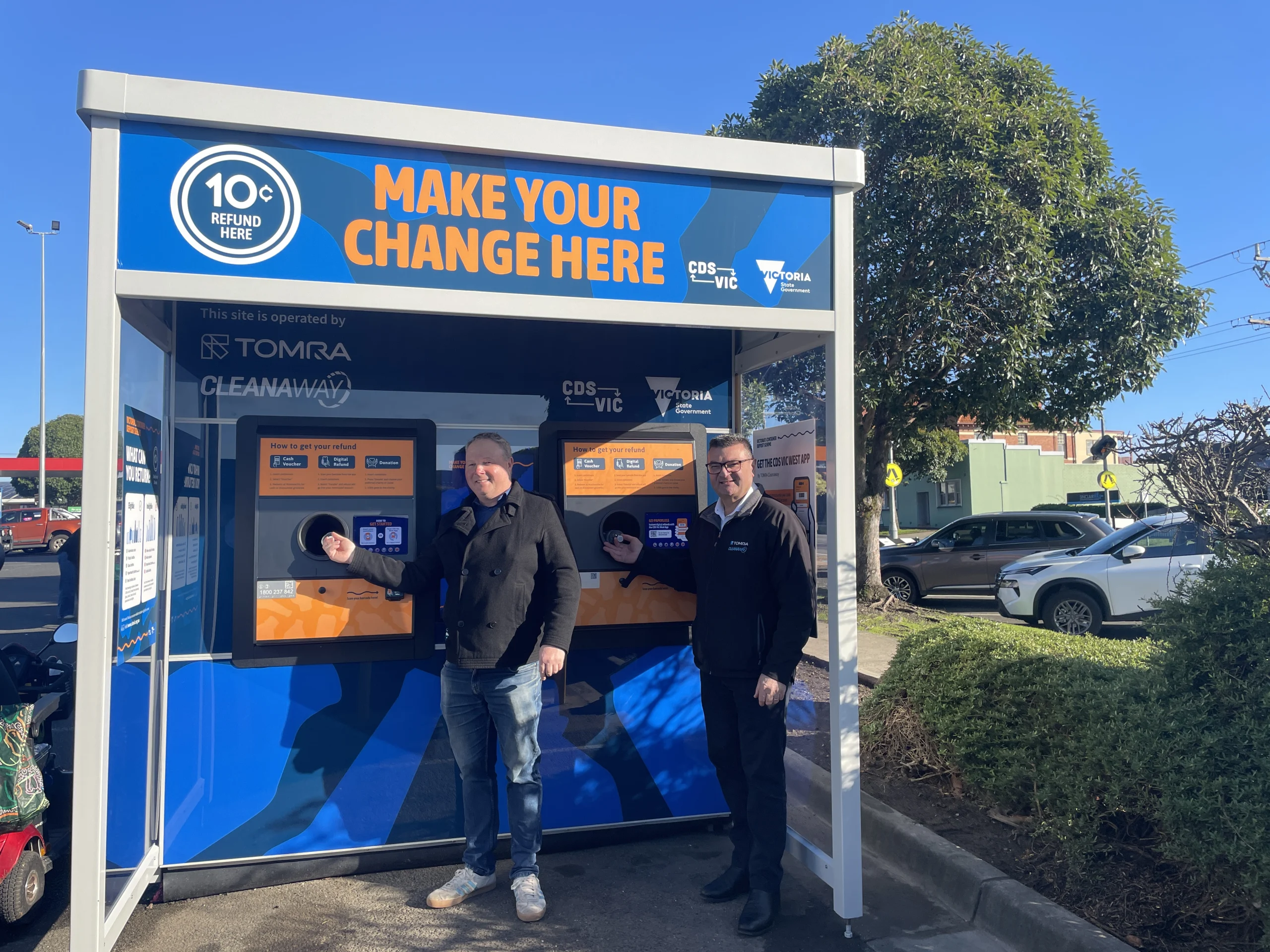 CDS Vic Colac Shopping Centre RVM opening - TOMRA Cleanaway Outdoor blue refund kiosk with bold orange text 'MAKE YOUR CHANGE HERE' and two men standing at the counter on a sunny day
