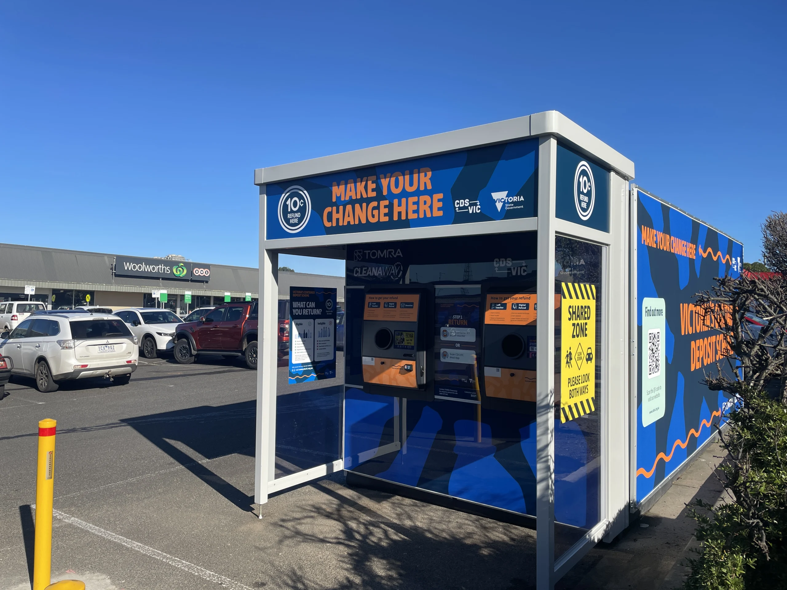Blue information kiosk in a parking lot with orange text reading 'MAKE YOUR CHANGE HERE' and circular '10c REFUND HERE' symbols; Woolworths store in background.