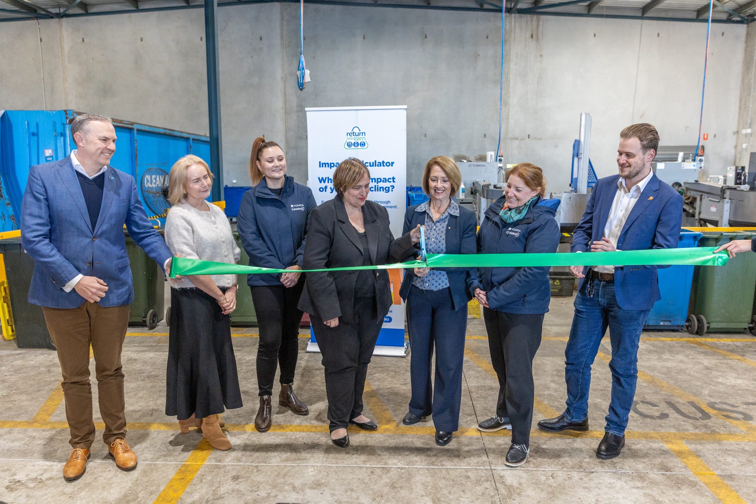Brent Murray TOMRA Danielle Smalley Exchange for Change Vesna Davidovic TOMRA Cleanaway TC Yolanda Saiz CEO Vinnies NSW Karen McKeown MP for Penrith Michelle Mandl TC Penrith Deputy Mayor Cr Gar - TOMRA Cleanaway Seven professionals in a factory ribbon-cutting ceremony, holding a bright green ribbon in front of a tall banner and industrial equipment behind them.