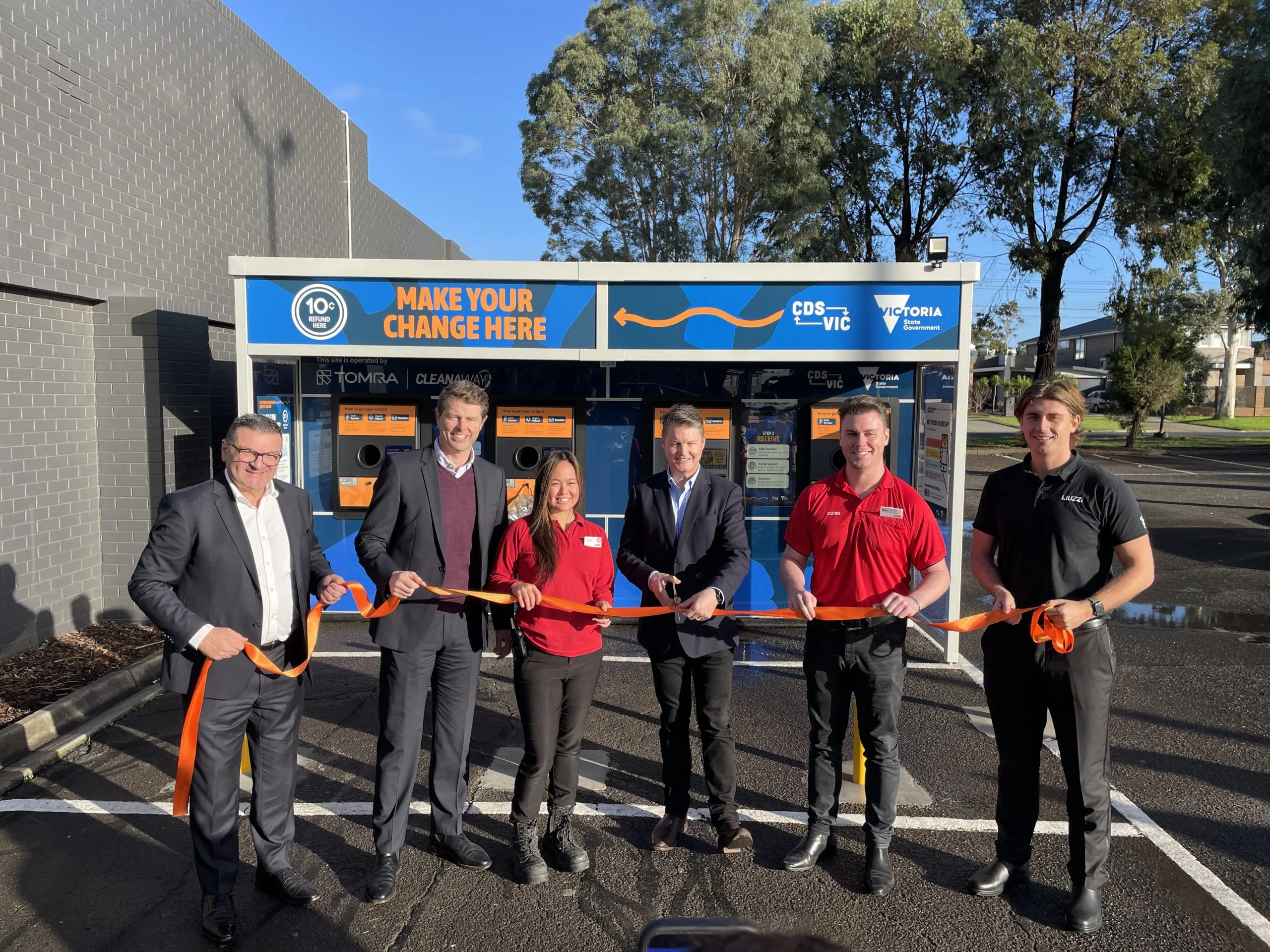 Six people stand in a line, cutting an orange ribbon in front of a blue, government-themed information booth during a ribbon-cutting ceremony.