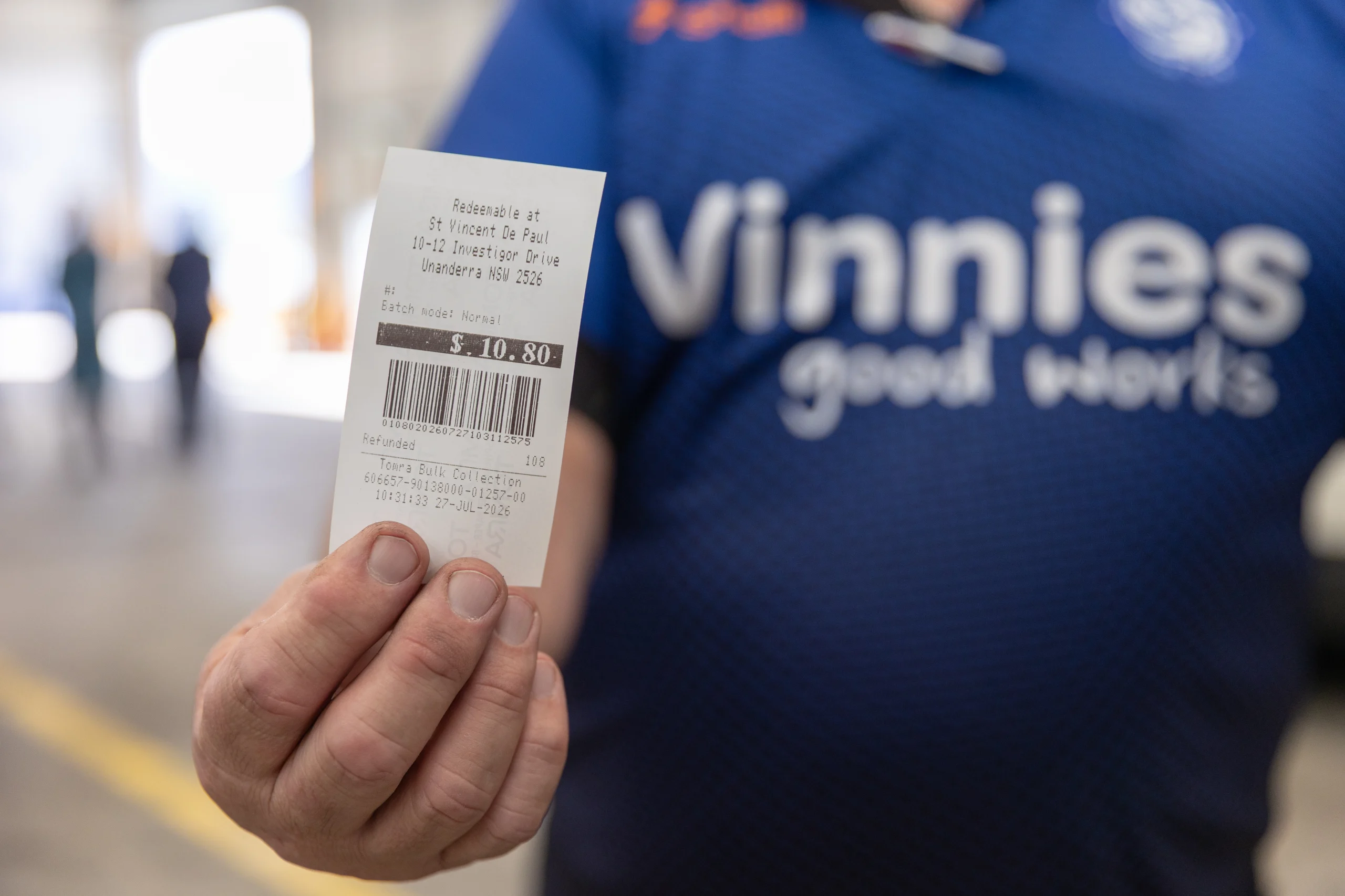 2026 Return and Earn Vinnie Unanderra TOMRA Cleanaway (2) - TOMRA Cleanaway Close-up of a hand holding a small receipt with a barcode; blurred background shows a blue shirt.1: