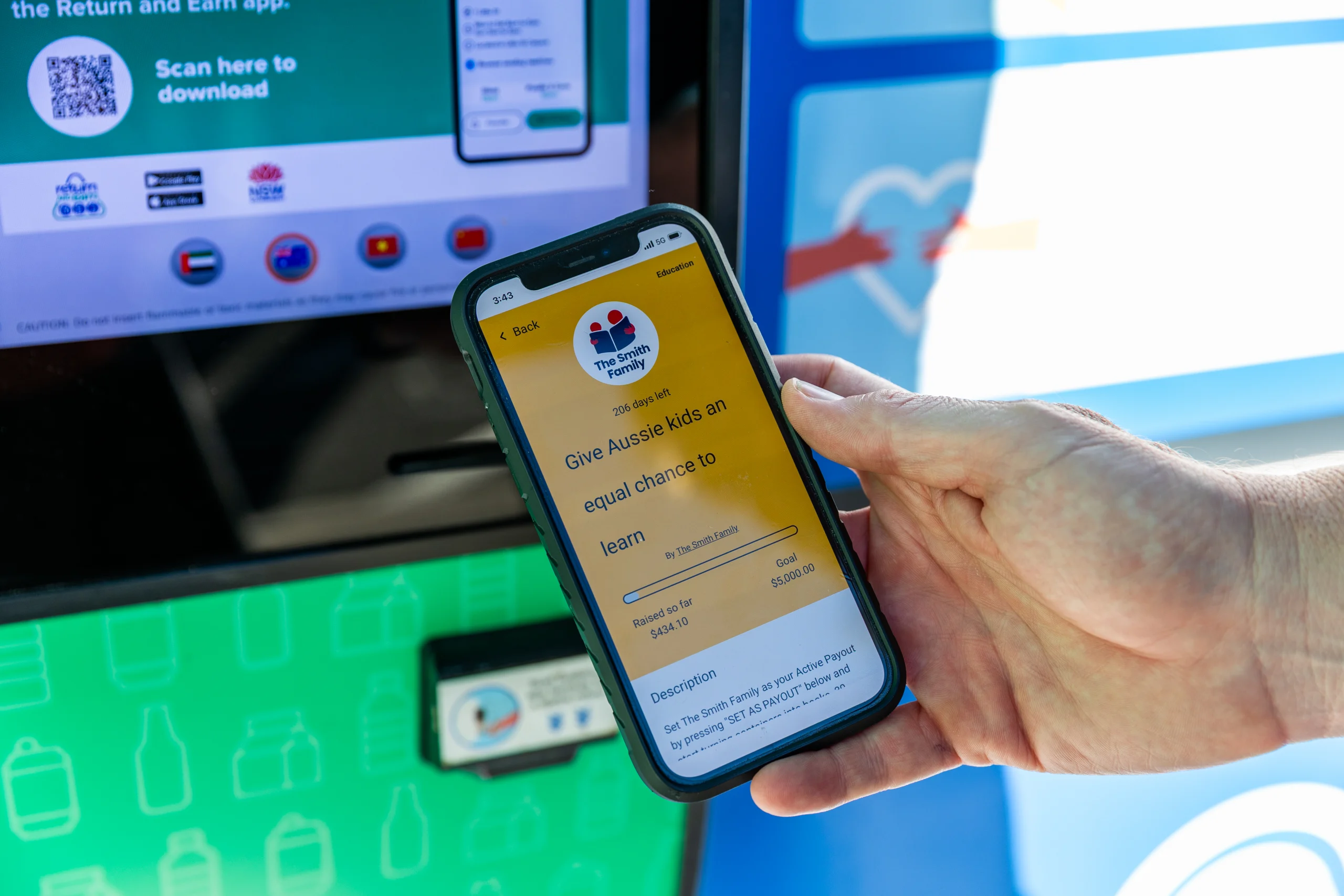 Person holding a smartphone showing The Smith Family donation screen, with a green kiosk and a QR code nearby? (informative)