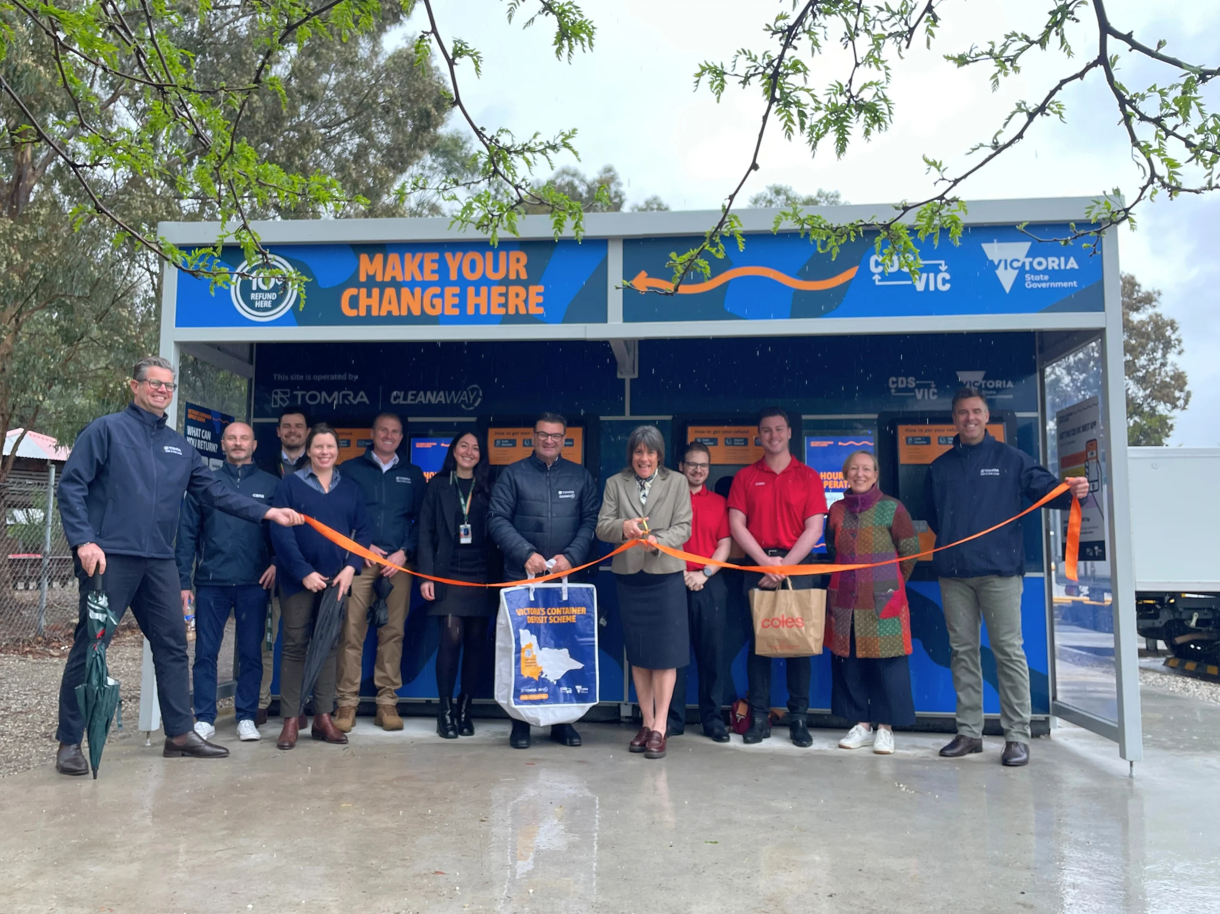 Group of people in jackets posing with orange ribbon in front of a blue 'MAKE YOUR CHANGE HERE' booth at a reverse vending machine opening event.