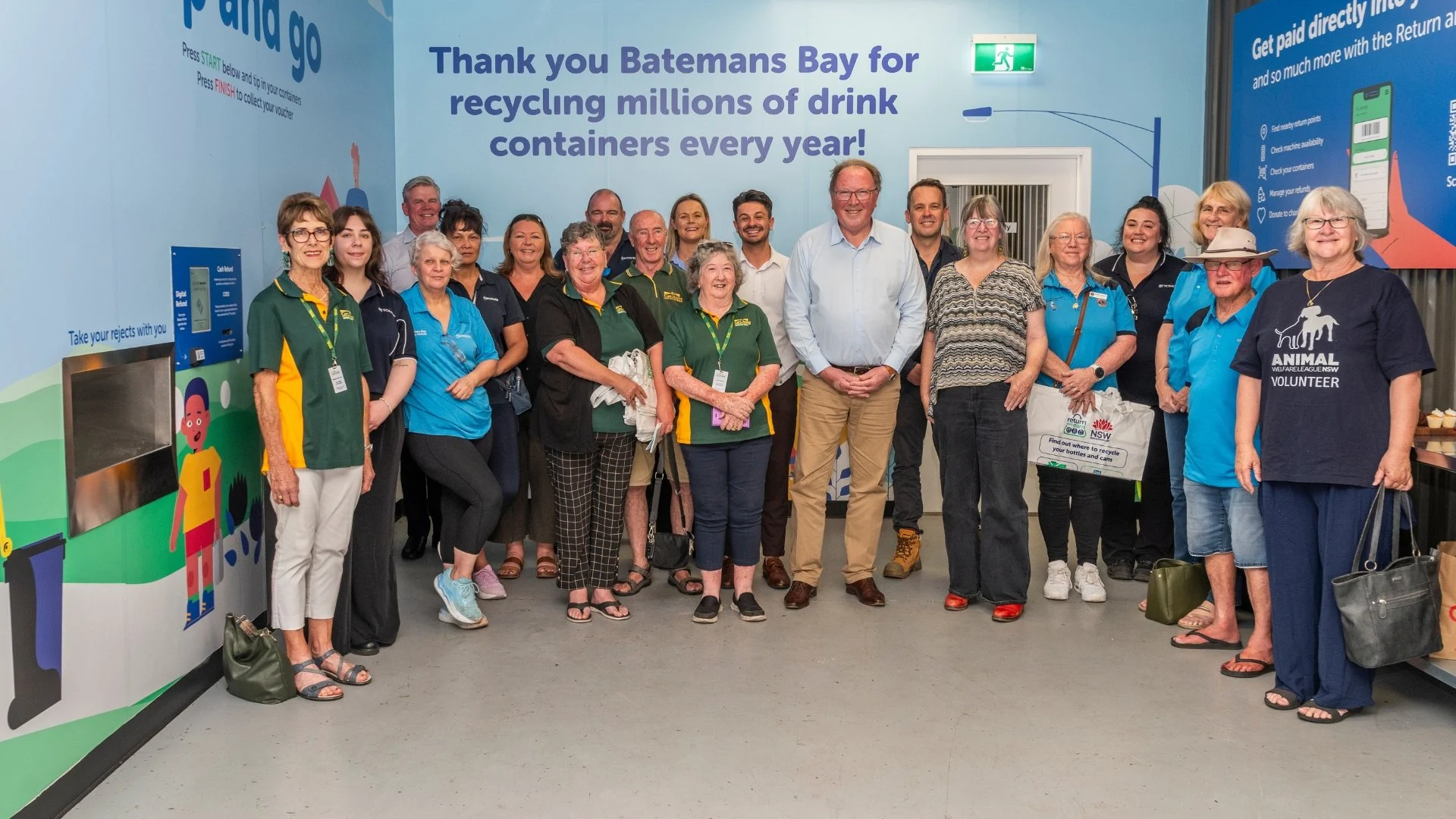 Group of volunteers and staff posing for a photo inside a recycling center, under a banner thanking Batemans Bay for recycling drink containers.