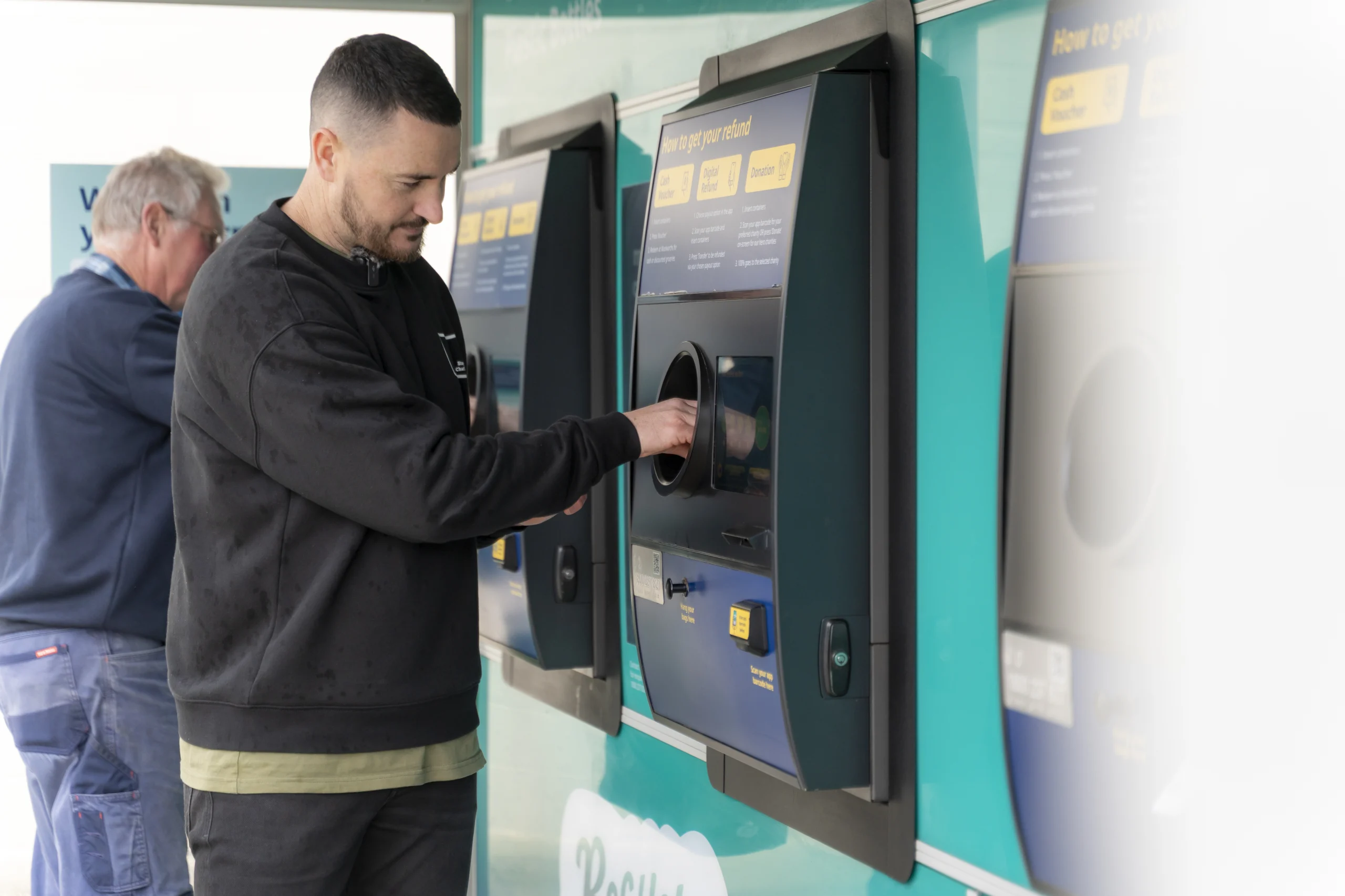 Man using a large self-service payment kiosk at a teal storefront, with another person in the background.