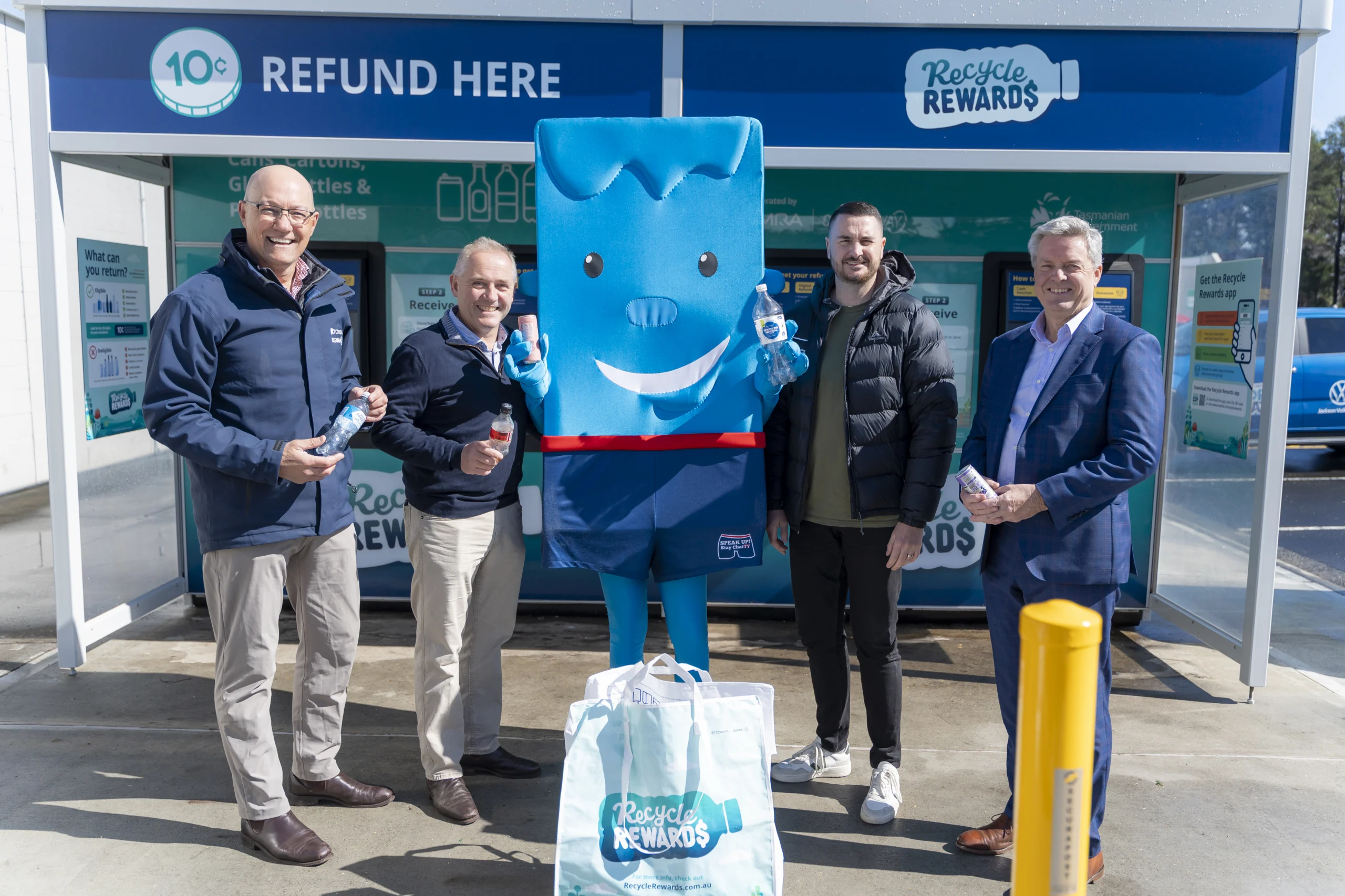 Four men in smart casual and jackets pose with a large blue recycling mascot outside a 'Recycle Rewards' booth, holding bottles and bags nearby as part of a recycling event.