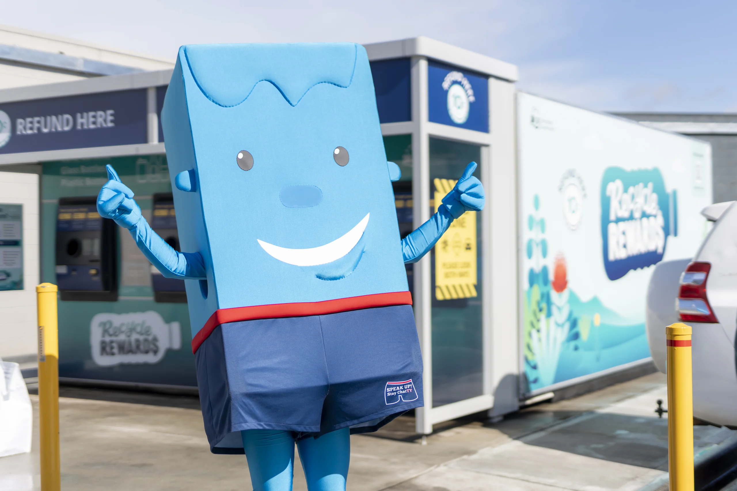 Blue square mascot with a friendly smile and raised thumbs stands outside a storefront, near a 'Refund Here' sign.